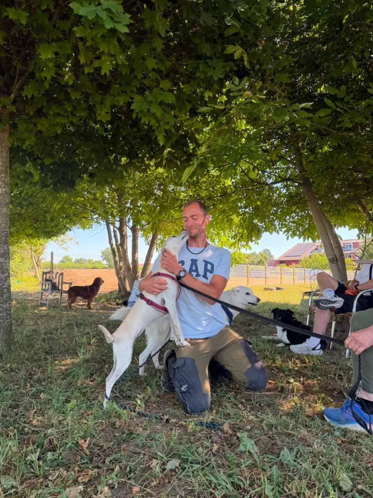 Robin Tast kniet im Schatten eines Baumes, ein weißer Hund springt an Robin hoch und gibt einen Kuss.